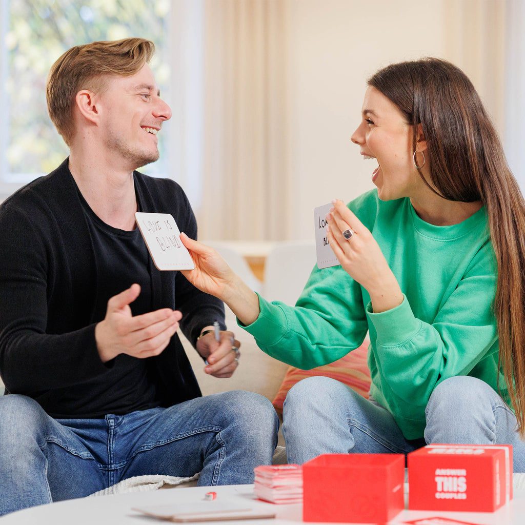 A happy couple playing a relationship card game in a cozy living room. The man, wearing a black sweater and jeans, laughs while holding a card that says 'Love is Blind.' The woman, dressed in a green sweatshirt and jeans, excitedly reacts while holding a similar card. A red game box labeled 'ANWSER THIS - COUPLES' sits on the table in front of them, along with a stack of cards. The warm and inviting indoor setting suggests fun, bonding, and engaging conversation through the game.